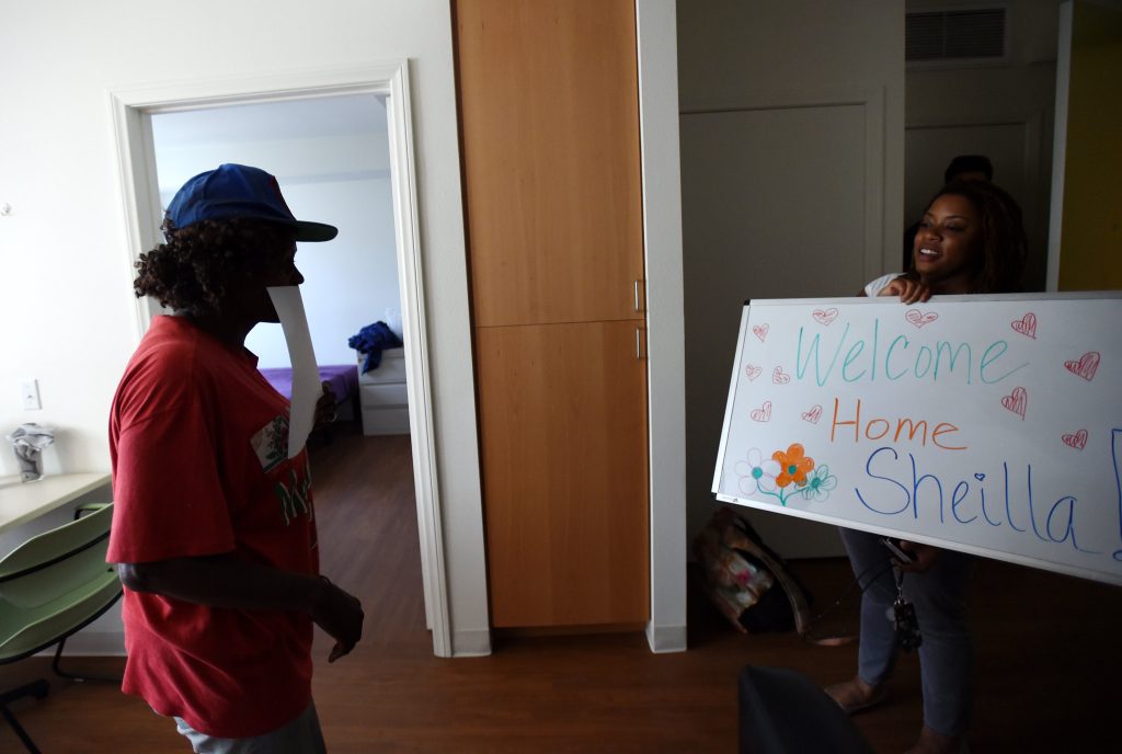 A woman holds a sign that says “Welcome Home Sheilla” inside an apartment.
