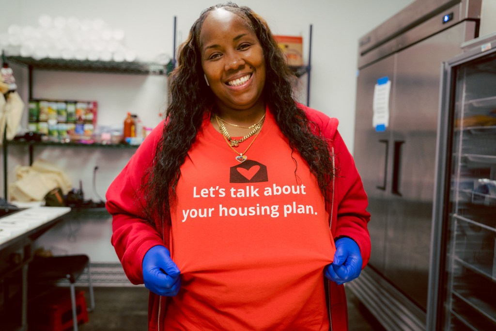 Smiling woman in red shirt that reads: “Let’s talk about your housing plan.”