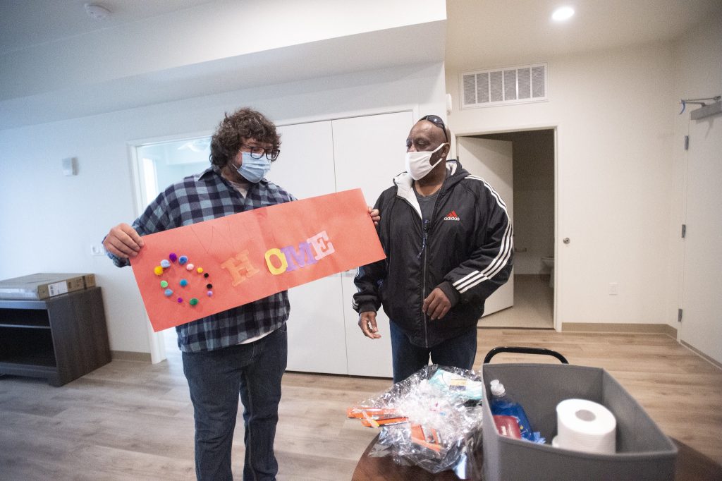 Two men stand indoors holding a red “Welcome Home” sign.