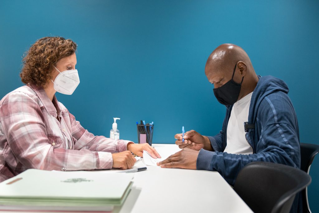 wo people wearing masks sit at a table while one signs a document.