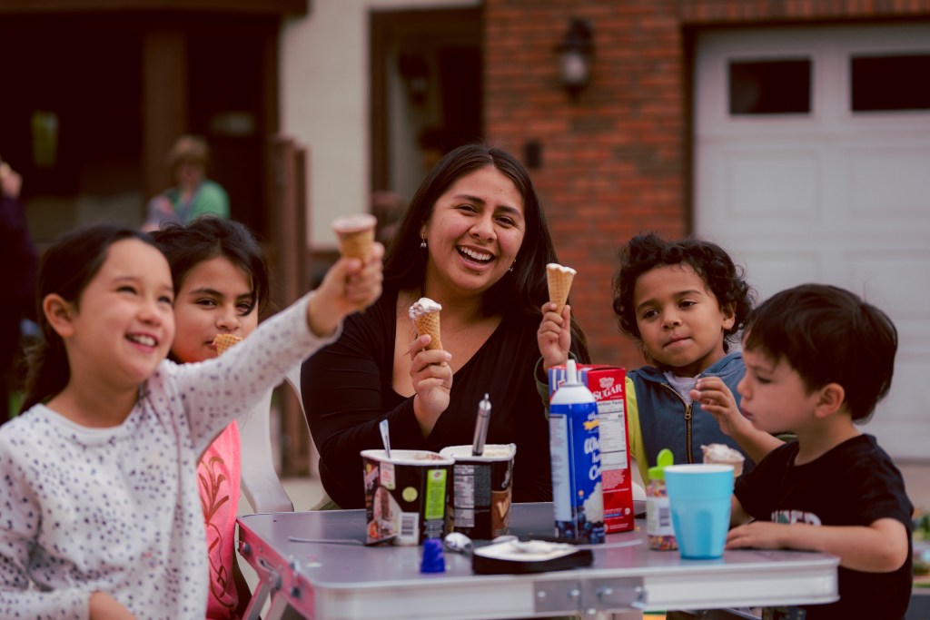 Smiling woman and children sit at a table enjoying ice cream cones in a neighborhood setting.