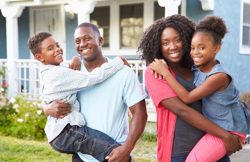 Smiling African-American parents each hold a child in front of a house with a white porch.