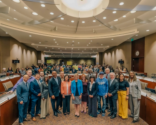 Team smiling in a large meeting room with an audience seated behind them.