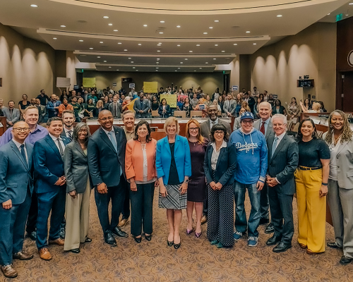 Team smiling in a large meeting room with an audience seated behind them.